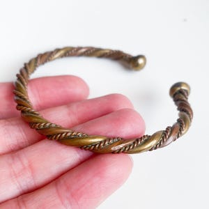Può includere: Un bracciale a polsino color bronzo con un design a corda intrecciata. Il bracciale aperto ha una sfera arrotondata a ciascuna estremità. Il metallo ha una miscela di tonalità oro e rame.
