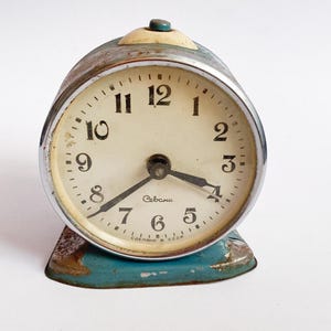 Pode incluir: Despertador vintage de estilo retro com mostrador creme, algarismos pretos e ponteiros pretos. O relógio tem uma caixa de metal prateado, uma base azul e um pequeno botão na parte superior. O mostrador mostra "Севани" e "СДЕЛАНО В СССР".