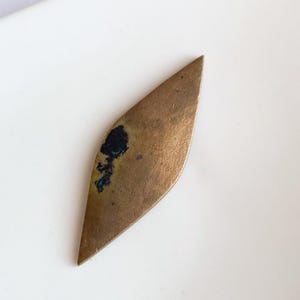 Op de afbeelding: Een vintage, bladvormig messing object met een aangetaste, verouderde uitstraling. Het messing heeft een warme, gouden tint met donkere oxidatievlekken, wat karakter toevoegt. De langwerpige vorm loopt aan beide uiteinden taps toe.