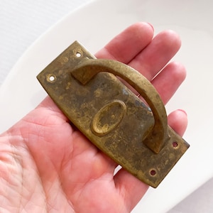May include: A vintage brass drawer pull with a rectangular backplate and a curved handle. The pull has an oval design in the center and three screw holes on each end. The brass has a tarnished, aged appearance.