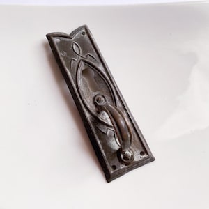 Könnte beinhalten: Antiker Türgriff in Bronzetönen mit Schlüsselloch und gebogenem Griff. Die rechteckige Platte weist dekorative, blütenartige Designs und kleine Löcher zur Montage auf. Ein Hardware-Teil im Vintage-Stil.