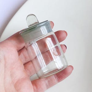 Puede incluir: Una botella de laboratorio de vidrio transparente con un tapón de vidrio. La botella tiene un cuerpo cilíndrico y un cuello acampanado. El tapón tiene un pequeño asa. La botella se sostiene en una mano.