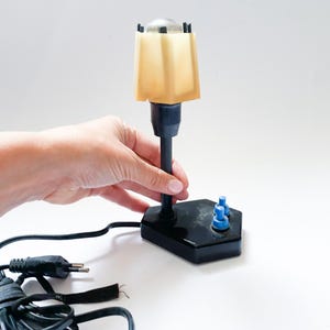 May include: A vintage desk lamp with a black base and stem, topped with a tan, square lampshade. The lamp has two blue buttons and a power cord. The lamp is held by a hand.