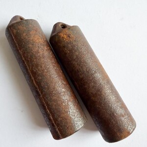 May include: Two cylindrical metal weights with a dark, oxidised, and rusted brown surface texture. Each object features a rounded top end with a small, circular hole drilled through the centre, designed for attachment or suspension purposes.