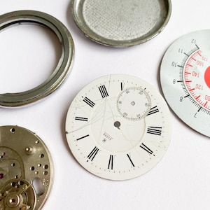 Op de afbeelding: Een verzameling vintage horlogeonderdelen, waaronder een gebarsten witte wijzerplaat met zwarte Romeinse cijfers en een kleine subwijzerplaat. Andere onderdelen zijn een metalen bezel, een horlogekast en een rood-witte wijzerplaat.
