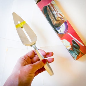 May include: A silver cake server with a yellow gummy sweet on the handle. The server is being held in a hand with red nail polish.