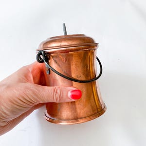 Może przedstawiać: Małe wiaderko w kolorze miedzi z pokrywką i czarnym uchwytem. Wiaderko ma cylindryczny kształt z lekko rozszerzoną podstawą. Mały, ciemny metalowy uchwyt jest przymocowany do pokrywy. Wiaderko trzymane jest w dłoni z czerwonym lakierem do paznokci.