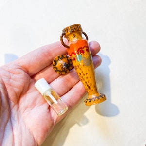 Könnte beinhalten: Eine kleine, holzerne Parfümflasche in Form einer Vase mit einem orangefarbenen, roten und blauen Design. Die Flasche hat einen kleinen, runden, hölzernen Deckel mit einem kupfernen Griff. Ein klares Glasfläschchen mit einem weißen Verschluss ist ebenfalls abgebildet.