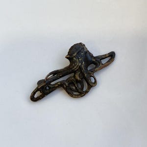Puede incluir: Un broche de metal de color bronce con un diseño intrincado de una figura estilizada con líneas fluidas.