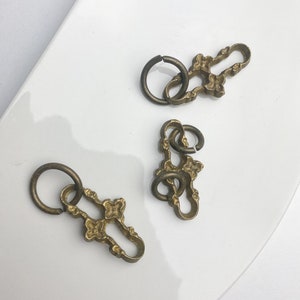 Puede incluir: Tres eslabones de cadena decorativos de metal dorado con diseños florales ornamentados. Cada eslabón tiene una abertura circular y está conectado al siguiente eslabón por un anillo circular más pequeño.