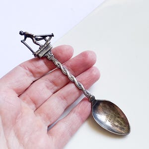 May include: Silver souvenir spoon with a twisted handle and a figure of a person on a starting block. The spoon bowl has the text "SOUVENIR FROM NORWAY".