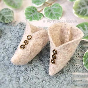 Puede incluir: Un par de zapatillas de fieltro beige con detalles de botones dorados. Las zapatillas están sobre una superficie texturizada azul y blanca.