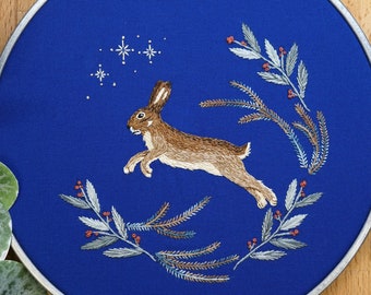 May include: A circular embroidery hoop containing blue fabric with a hand-stitched brown rabbit leaping through the air. The design features white embroidered stars above the rabbit and two curved branches with small red berries framing the scene.