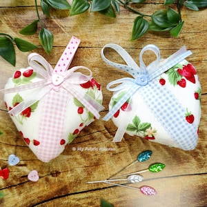 May include: Two heart-shaped fabric pincushions with a strawberry print and gingham ribbon accents. The pincushions are filled with stuffing and have buttons in the centre. The pincushions are on a wooden surface with green leaves and sewing pins.