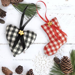 May include: Two handmade Christmas ornaments, one in the shape of a heart and one in the shape of a stocking. The heart ornament is black and white gingham with a black ribbon and a yellow star button. The stocking ornament is red and white gingham with a red ribbon and a yellow star button. Both ornaments are hanging from a white wooden background.