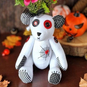 May include: A white handmade teddy bear with black and white polka dot fabric accents. The bear has red button eyes and a spiderweb embroidered on its chest.