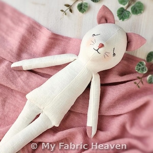 Könnte beinhalten: Eine cremefarbene Stoffpuppe in Katzenform mit rosa Ohren und rosa Akzenten an den Pfoten. Die Puppe hat gestickte geschlossene Augen, eine kleine rosa Nase und Schnurrhaare. Der Text My Fabric Heaven ist auf der Puppe zu sehen.