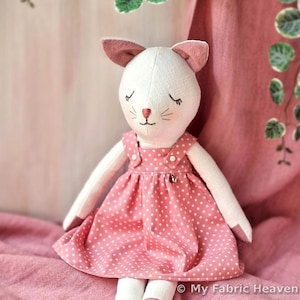 May include: A handmade stuffed cat doll with a white head, pink ears, and a pink polka dot dress. The doll has closed eyes and a stitched face. The doll is sitting on a pink surface.