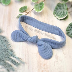 May include: A light blue fabric headband with a bow tied in the center. The headband is made of a soft, textured fabric and is perfect for newborns and babies.