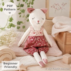 Könnte beinhalten: Ein Stofftier in Form einer Katze aus cremeweißem Stoff mit gestickten geschlossenen Augen und einer rosa Nase. Die Katze trägt ein Oberteil mit Blumenmuster in Creme und Rot sowie eine passende rote, dazu passende rote Blumen-Pluderhosen. Die Ohren haben rosa Stoffeinsätze.