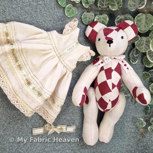 May include: A handmade teddy bear with a red and white checkered pattern on its torso, ears, and feet, alongside a cream-colored dress with gold trim and lace details. A matching headband with a bow is also present. The items are likely for a child.
