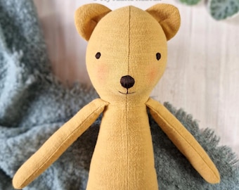 May include: A mustard yellow fabric bear doll with embroidered dark brown eyes and a nose. The bear features soft pink blush on its cheeks and a vertical seam running down the center of its face. The text My Fabric Heaven is visible above.