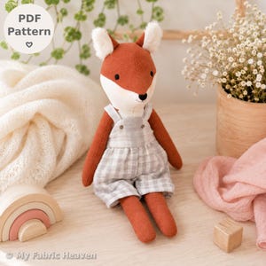 Puede incluir: Un peluche de zorro con pelaje color óxido, marcas faciales blancas y orejas esponjosas, que lleva un peto de cuadros grises y blancos. El zorro tiene ojos y nariz bordados en negro. Una etiqueta circular en la esquina dice PDF Pattern.