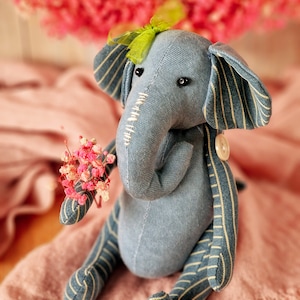 May include: A blue and white striped fabric elephant toy holding a bouquet of pink and white flowers. The elephant has a green bow on its head and a button on its ear.