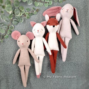 Könnte beinhalten: Vier Stoffpuppen, darunter eine beige Maus mit rosa Ohren, eine weiße Katze mit rosa Ohren, ein rostfarbener Fuchs mit weißen Akzenten und ein blassrosa Kaninchen mit langen Ohren. Der Text My Fabric Heaven ist unten rechts aufgedruckt.