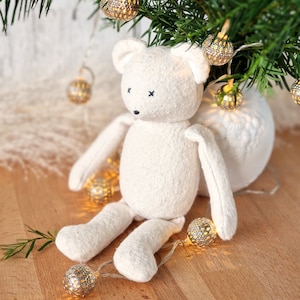 Könnte beinhalten: Ein weißer Plüschteddybär mit schwarzen gestickten Augen sitzt auf einer Holzoberfläche, umgeben von einer Lichterkette mit goldenen Weihnachtslichtern.
