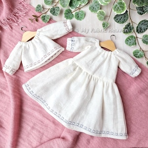 Puede incluir: Una blusa blanca de manga larga y un vestido blanco a juego, ambos con un delicado patrón de corazones bordados a lo largo del dobladillo y los puños. Cada prenda se exhibe en una percha de madera, mostrando una tela texturizada.