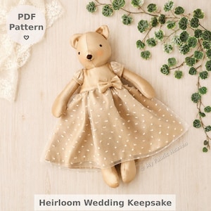 May include: A plush teddy bear wearing a beige dress with white polka dots and a bow. The bear has a stitched nose and a sweet expression. The text "Heirloom Wedding Keepsake" is at the bottom.