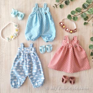 May include: A collection of doll clothes, including a blue polka dot romper, a blue cloud-print jumpsuit, and a pink polka dot dress. Accessories include shoes and floral headbands. The items are arranged on a light wood surface.