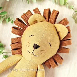 May include: A plush lion toy made of mustard yellow fabric with a dark brown stitched nose and mouth. The mane consists of looped dark brown fabric strips. The text My Fabric Heaven is printed in white at the bottom of the lion's chest area.