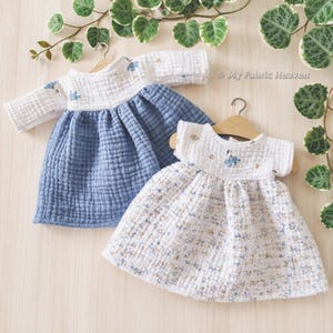 Pode incluir: Dois vestidos de musselina em cabides de madeira. O vestido da esquerda tem mangas compridas, um corpete branco com detalhes florais azuis e uma saia azul sólida. O vestido da direita é sem mangas, com um corpete branco bordado com flores azuis.