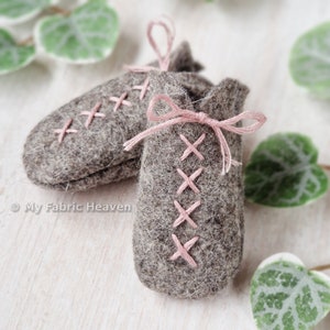 May include: A pair of grey felt baby booties with pink laces and a criss-cross stitch pattern. The booties are tied with a bow.