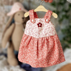 May include: A red and white floral print dress with a gathered skirt and straps. The dress has a small flower embellishment on the bodice.