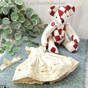 May include: A handmade teddy bear with a red and white chequered pattern, a cream-coloured dress with lace trim, and a matching ribbon. The bear has a red button and a white ribbon bow. The dress is cream-coloured with gold accents.