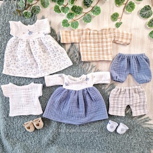 May include: A selection of doll clothing in different styles and colours. The set includes dresses, shirts, and shorts in white, blue, and tan. Two pairs of doll shoes are also included. The clothes are made of a textured fabric.