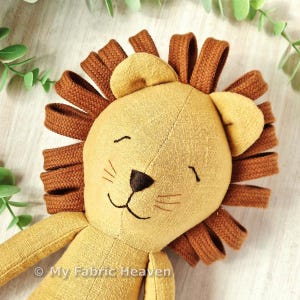 May include: A plush lion toy with a light brown body and a dark brown mane. The lion has a smiling face with embroidered details, including a black nose and whiskers. The toy is made of soft fabric and is designed for children.