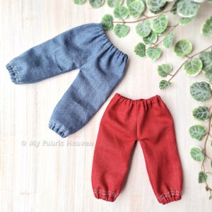 May include: Two pairs of doll trousers, one blue denim and one red. Both pairs have white heart-shaped stitching around the hems.