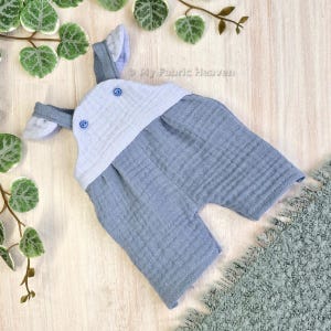 Puede incluir: Un mono de bebé azul claro con una parte superior azul claro y blanca y dos pequeñas orejas en los hombros. El mono tiene dos botones azules en las correas. "My Fabric Heaven" está escrito en la tela.