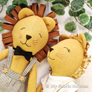 May include: Two handmade lion plush toys. The lions have yellow bodies, brown ribbon manes, and embroidered facial features. One lion wears a black bow tie and striped overalls. The other lion wears a white shirt.