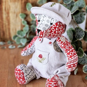 May include: A handmade patchwork teddy bear wearing a white cap and overalls with a red and white pattern. The overalls have a small embroidered mushroom design.
