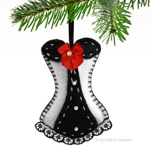 May include: A black and white felt Christmas ornament shaped like a corset with a red bow and white pearl accents. The ornament is hanging from a green pine branch.