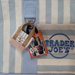 May include: A light blue and cream striped canvas tote bag featuring the embroidered blue text Trader Joe's. Attached to a metal D-ring is a miniature tote bag charm and a rectangular tag displaying a cupcake, a bottle, and the text I love TJ.