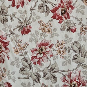May include: Floral fabric with a repeating pattern of large red and white flowers, smaller clusters of peach-colored blossoms, and gray leaves on a cream background. The fabric is measured with a yellow measuring tape.