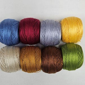 May include: Eight small balls of embroidery thread arranged in two rows. The colors include blue, deep red, light gray, yellow, beige, bronze, dark brown, and green. Each ball features a smooth, shiny texture with visible thread winding.