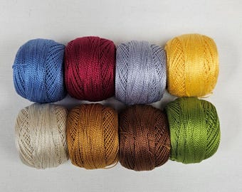 May include: Eight small balls of embroidery thread arranged in two rows. The colors include blue, deep red, light gray, yellow, beige, bronze, dark brown, and green. Each ball features a smooth, shiny texture with visible thread winding.