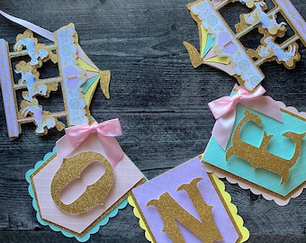 May include: A colorful banner with the word "ONE" in gold glitter letters. The banner is decorated with pastel pink, blue, and yellow paper cutouts of a carousel with horses. The banner is perfect for a first birthday party.